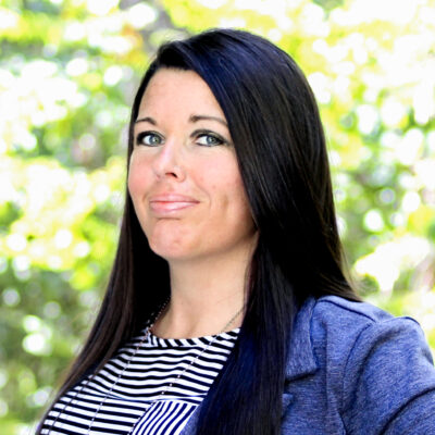 Amanda Hall wearing black and white striped shirt with light blue blazer in front of bright, outdoor, leafy background.