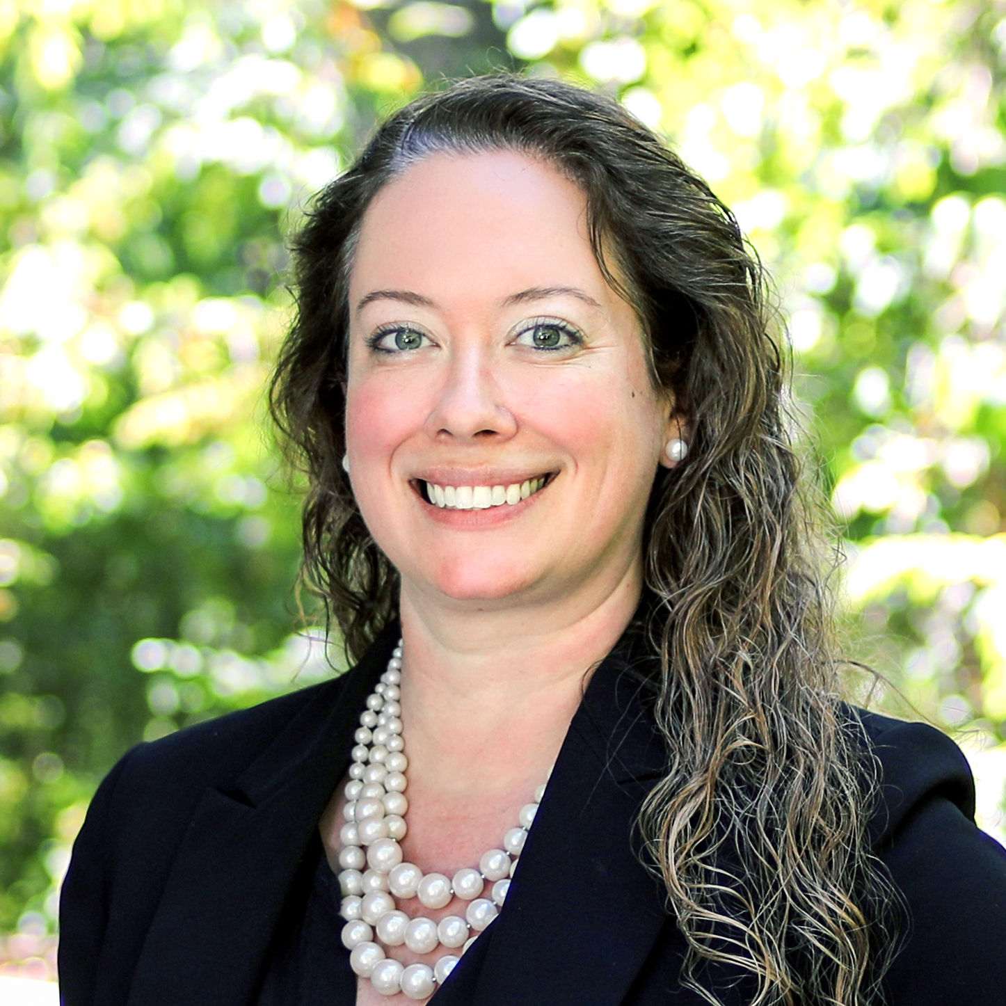 Erin Kennedy Startzman wearing black blazer and white pearls in front of bright, outdoor, leafy background.