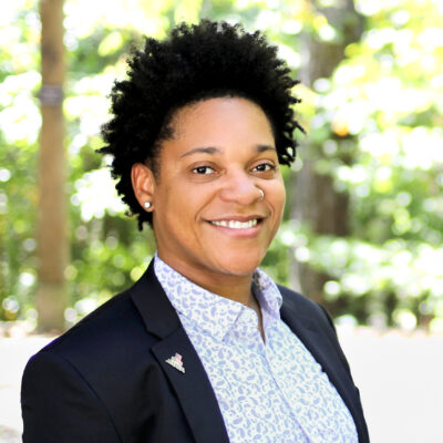 Keturah Herron wearing blue and white paisley button down with black blazer in front of bright, outdoor, leafy background.