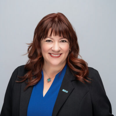 A smiling white woman with red hair wearing a blue top and black jacket.