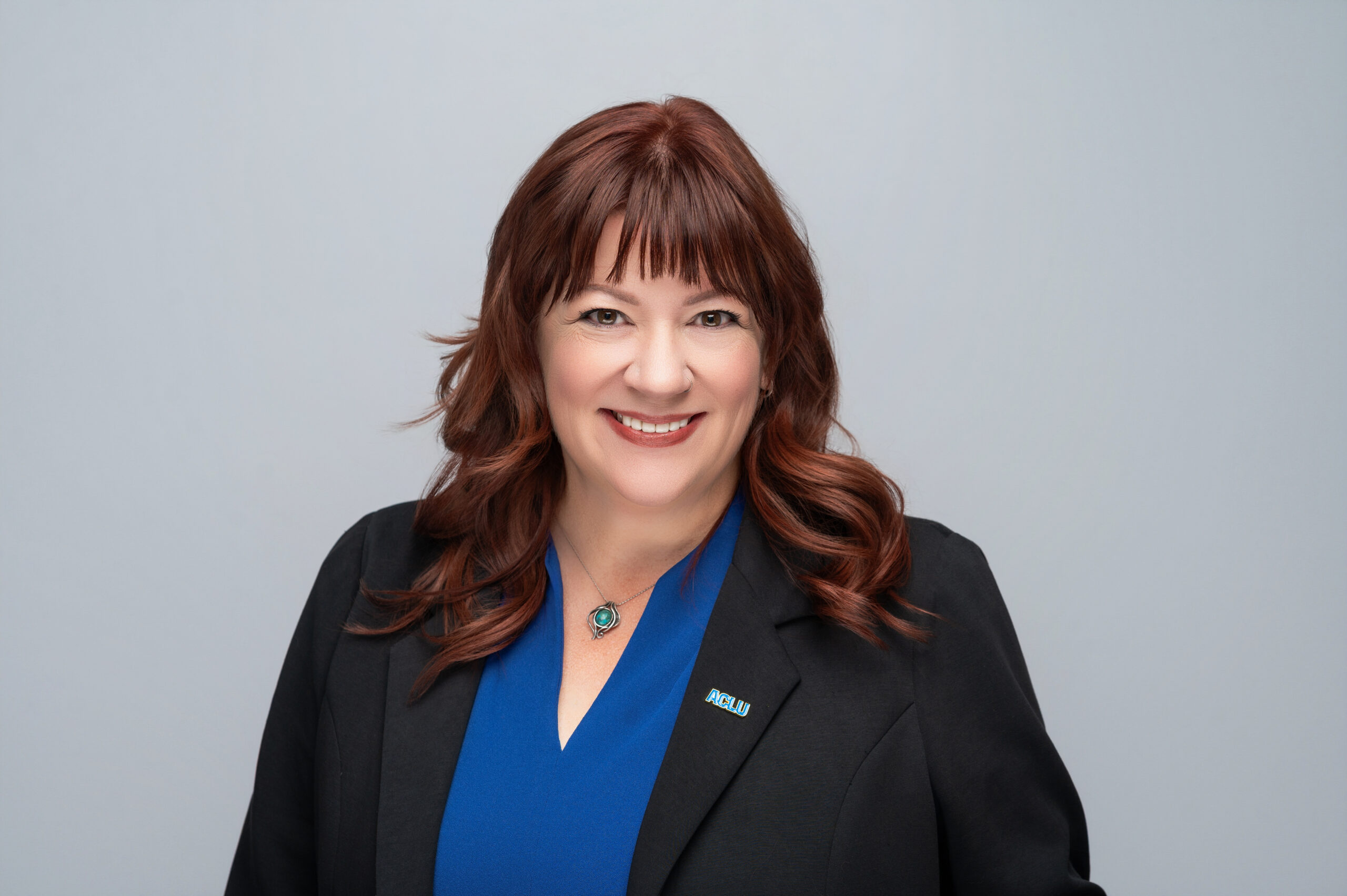 A smiling white woman with red hair wearing a blue top and black jacket.