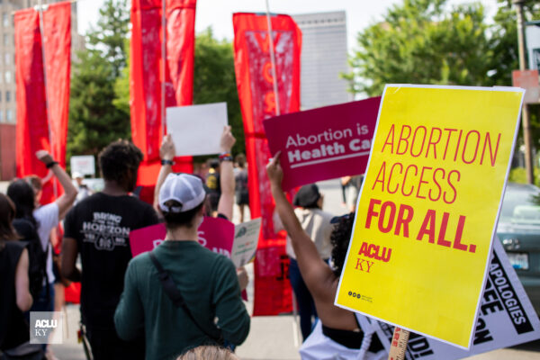 Protesters hold signs at rally in Louisville, KY. Yellow poster on right half reads, "Abortion access for all." Other signs are illegible.