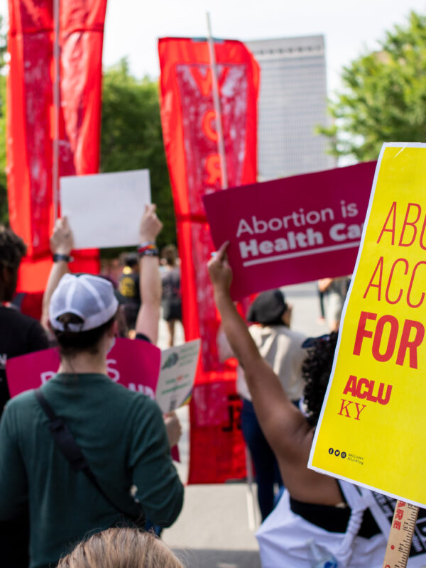 Protesters hold signs at rally in Louisville, KY. Yellow poster on right half reads, "Abortion access for all." Other signs are illegible.