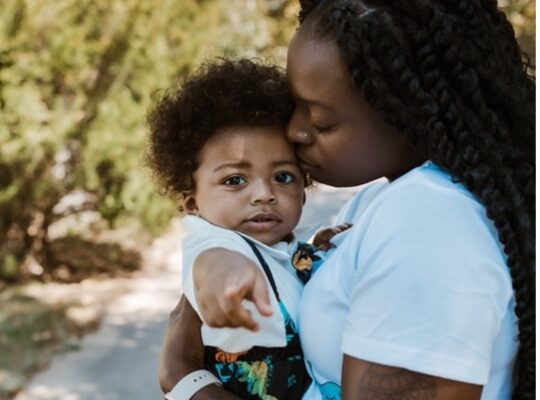 Katima holds her son, Mylo, as Mylo points to the camera and Katima kisses his forehead.