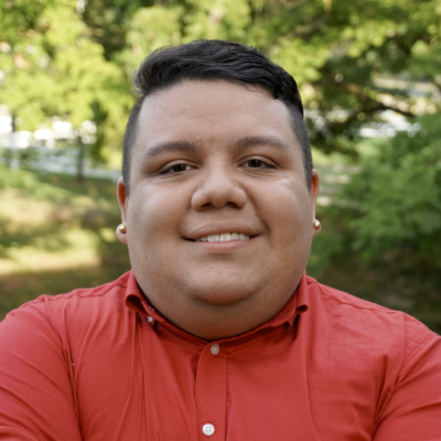 Omar Salinas-Chacón wearing dark t-shirt with like pink button down and white peals, standing in front of bright, outdoor, leafy background.