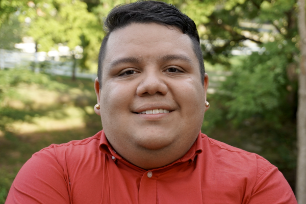 Omar Salinas-Chacón wearing dark t-shirt with like pink button down and white peals, standing in front of bright, outdoor, leafy background.