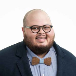 A smiling Latinx person with a short beard wearing glasses and a bow tie, with a light blue shirt and a dark blazer.