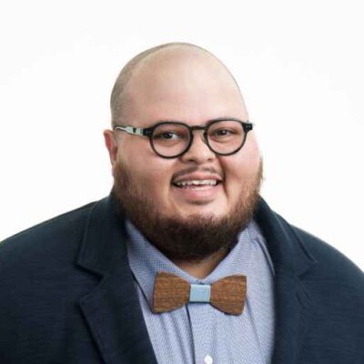 A smiling Latinx person with a short beard wearing glasses and a bow tie, with a light blue shirt and a dark blazer.