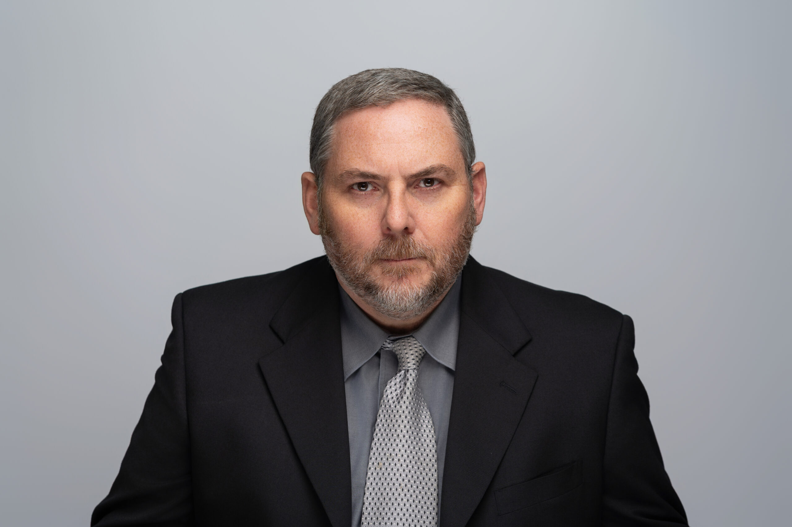 bill sharp, stern to camera, wearing a black suit coat and a grey shirt and tie
