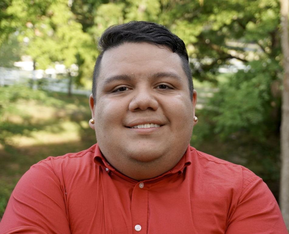 Omar Salinas-Chacón wearing dark t-shirt with like pink button down and white peals, standing in front of bright, outdoor, leafy background.