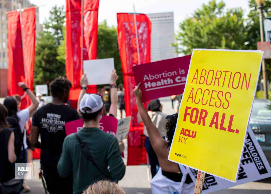 Protesters hold signs at rally in Louisville, KY. Yellow poster on right half reads, "Abortion access for all." Other signs are illegible.