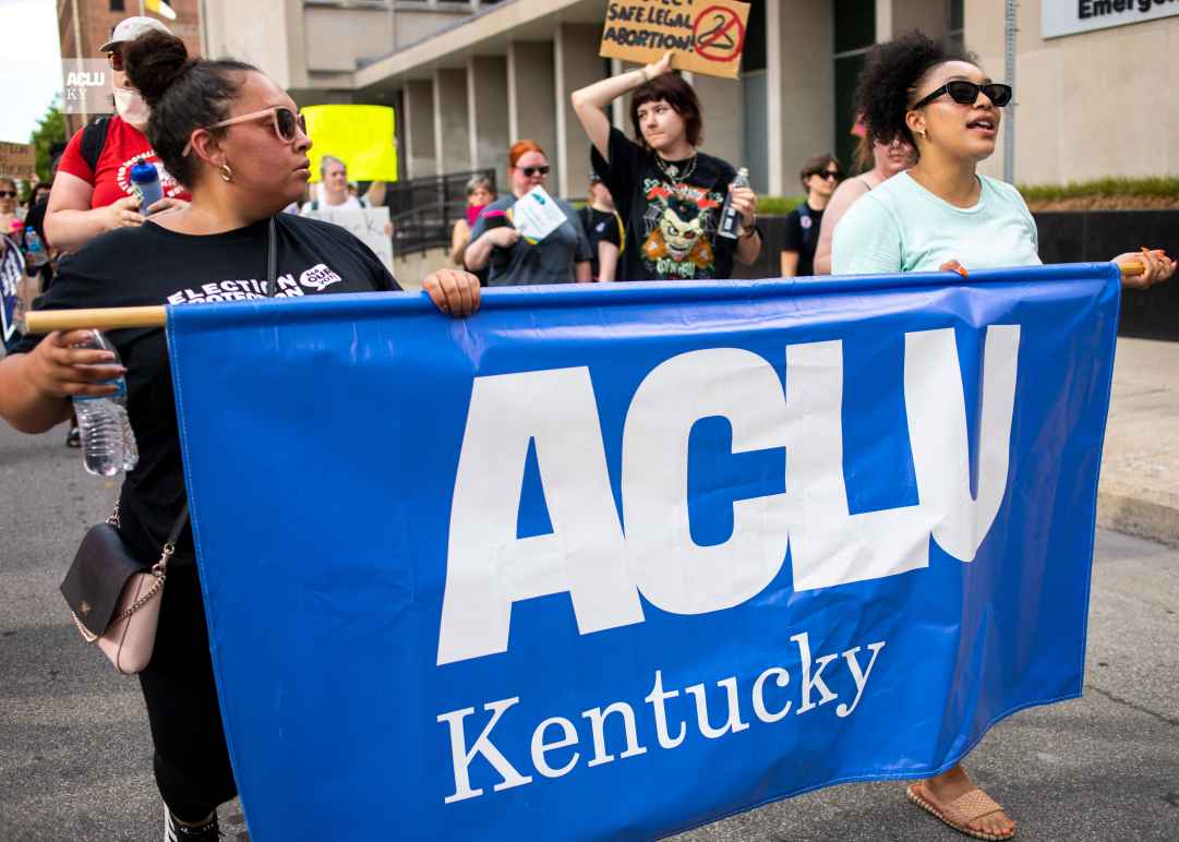 Two people carrying a blue ACLU of Kentucky banner during a march in streets in downtown Louisville KY